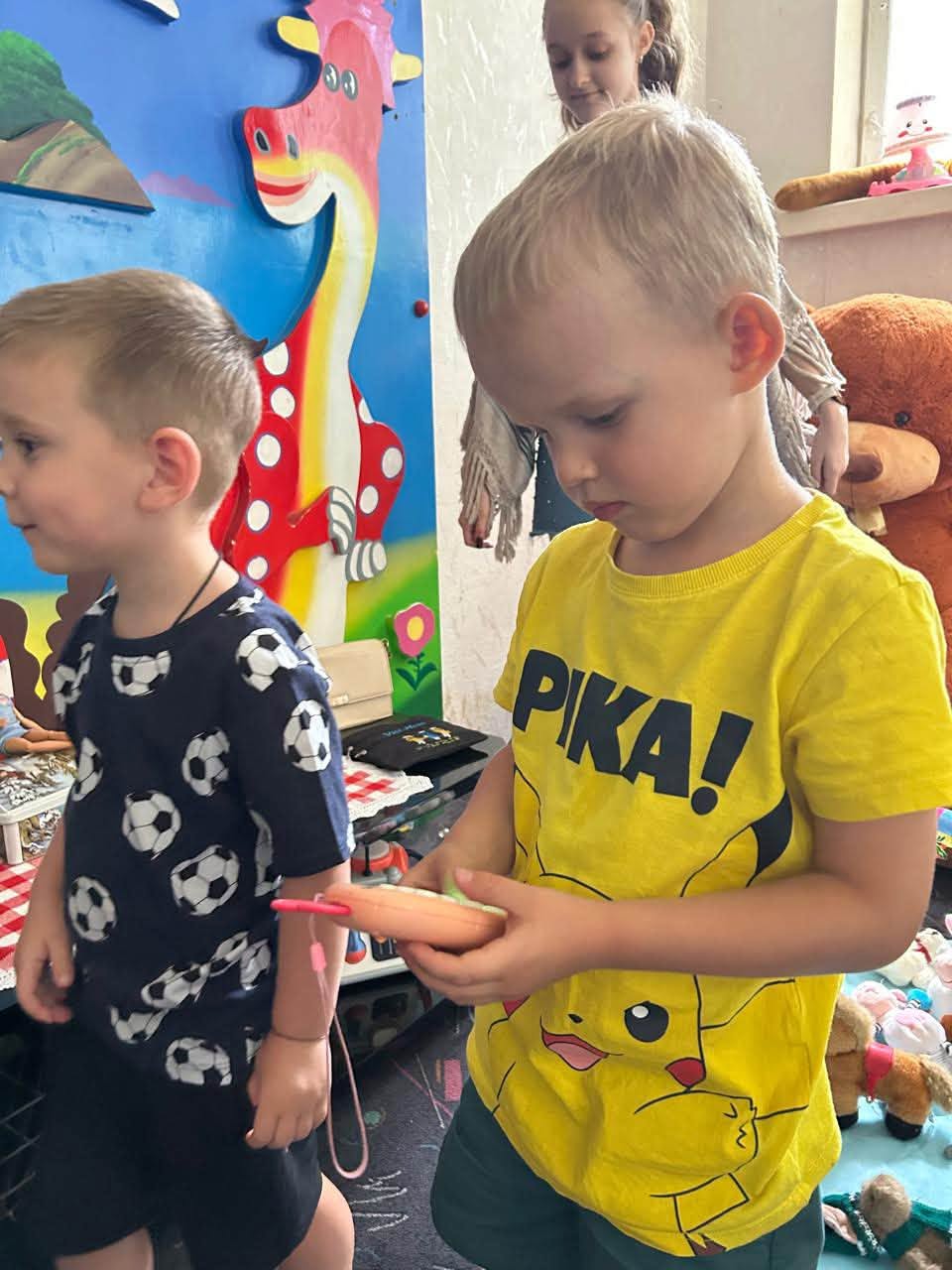 Хлопчик у яскраво-жовтій футболці з написом «PIKA!» та малюнком Пікачу тримає в руках рожеву дитячу іграшку у формі телефона з антеною. Поруч із ним стоїть інший хлопчик у темно-синій футболці з футбольними м'ячами. На задньому плані видно дівчинку-підлітка та яскраву ігрову стіну із зображенням червоно-жовтого динозавра й великі м'які іграшки.