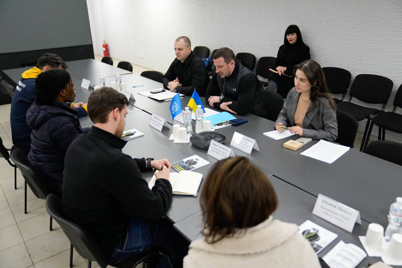 Група людей сиділа за довгим столом під час ділової зустрічі. На столі встановлені маленькі прапори України та ООН, а також таблички з іменами та блокноти.