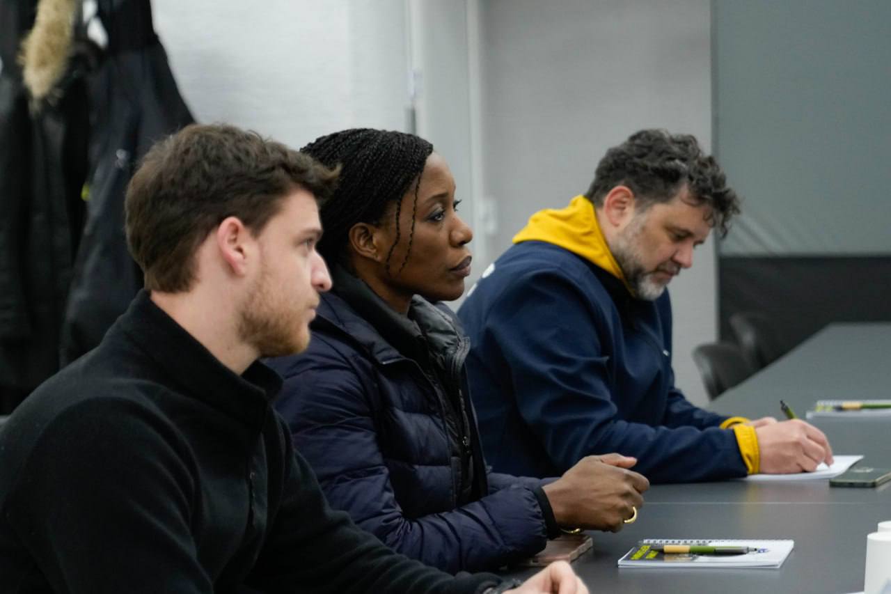 Троє учасників зустрічі крупним планом, які сидять за столом і зосереджено слухають або роблять записи у блокнотах під час офіційного заходу.