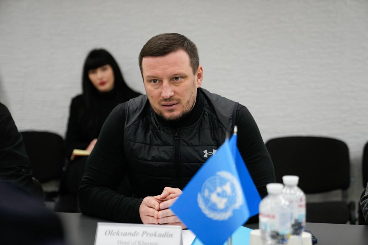 Учасник зустрічі крупним планом за столом, перед яким стоїть настільний прапор ООН та табличка з іменем. На фоні видно іншу особу та стіну з фактурною поверхнею.