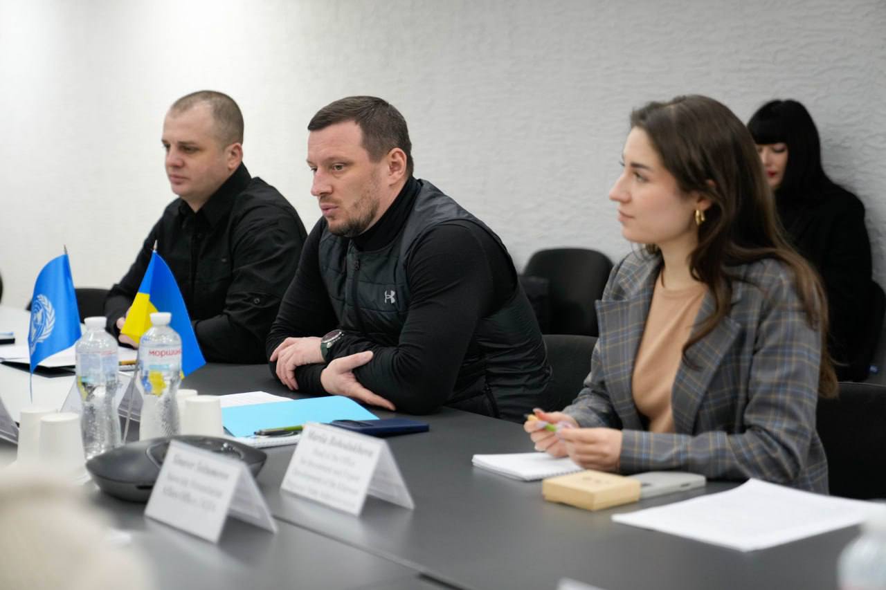 роє учасників зустрічі в напівпрофіль за столом, перед ними стоять настільні прапори та таблички з іменами.