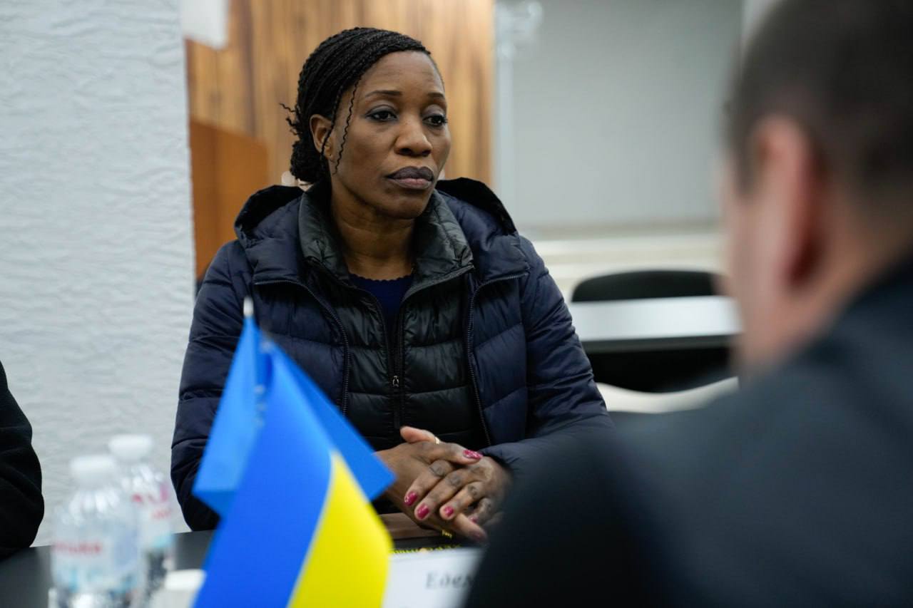 Жінка в темному верхньому одязі сидить за столом переговорів, склавши руки перед собою. На передньому плані видно розмиті силуети маленьких прапорів України та ООН, а також пляшки з водою.