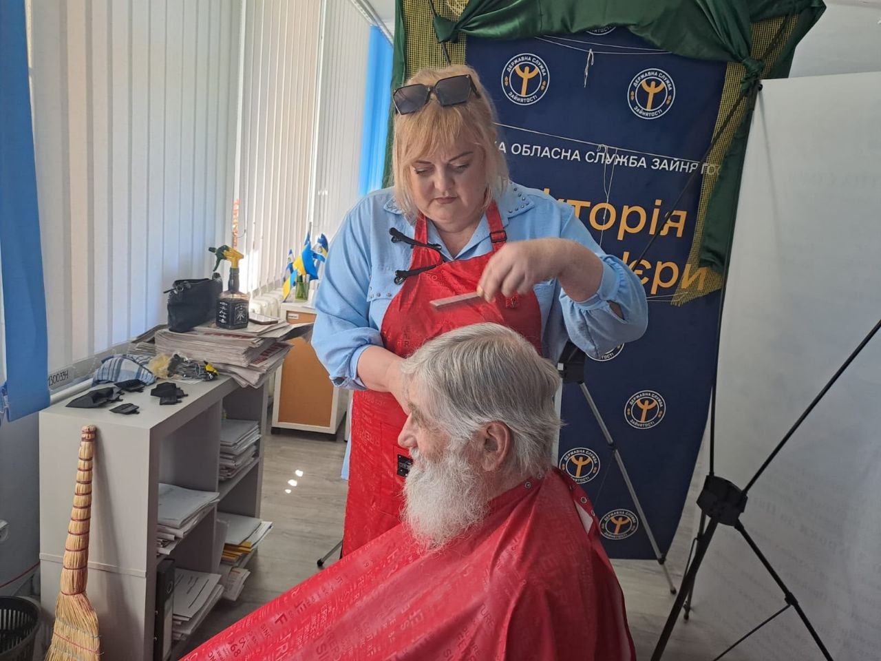 Жінка у білій блузці в чорний горошок стоїть біля білого фліпчарта та прикріплює магнітом деталі творчої аплікації. На аркуші зображено великий намальований плетений кошик із трьома паперовими соняшниками та зеленим листям. На жовтих пелюстках соняшників від руки написані побажання миру, здоров'я, спокою та перемоги. Унизу біля дошки стоїть червоний вогнегасник та розкладені маркери з губкою.