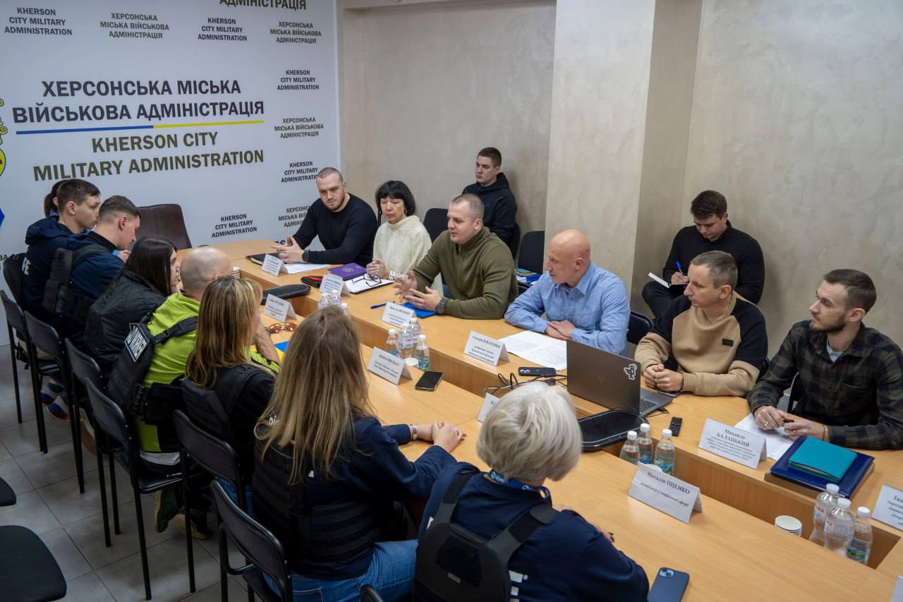 Нарада у приміщенні Херсонської міської військової адміністрації (МВА). Група представників адміністрації та гуманітарних організацій, деякі з яких у захисних жилетах, сидять за довгим столом. Чоловік у зеленій кофті активно виступає та жестикулює. На тлі видно повторюваний напис ХЕРСОНСЬКА МІСЬКА ВІЙСЬКОВА АДМІНІСТРАЦІЯ.