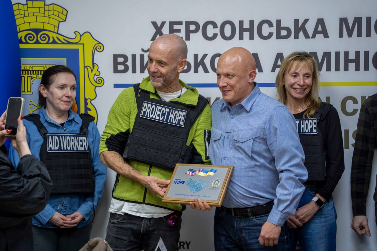 Два чоловіки посміхаються, тримаючи нагородну табличку з логотипами Project HOPE та прапорами України і США. Один чоловік одягнений у бронежилет із написом Project HOPE, інший – у синю сорочку. На тлі видно герб Херсона та напис ХЕРСОНСЬКА МІСЬКА ВІЙСЬКОВА АДМІНІСТРАЦІЯ.