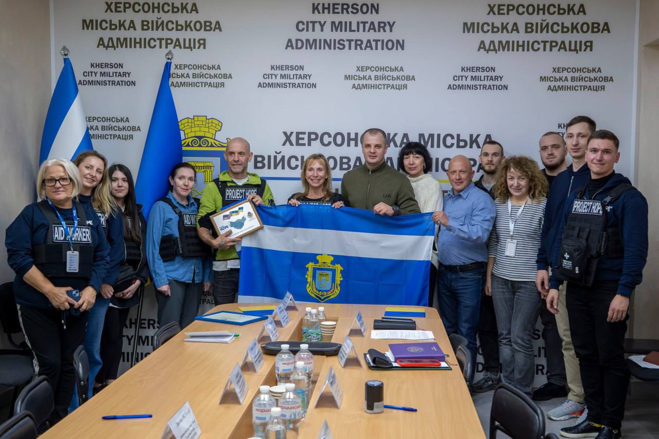 Група людей, включно з представниками військової адміністрації та міжнародних/гуманітарних організацій, стоїть у приміщенні Херсонської міської військової адміністрації. Вони тримають прапор Херсона та сертифікат подяки. На тлі видно повторюваний логотип ХЕРСОНСЬКА МІСЬКА ВІЙСЬКОВА АДМІНІСТРАЦІЯ та прапори України.