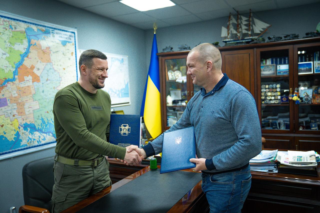 Олександр Прокудін та Дмитро Зорик тиснуть руки в робочому кабінеті, тримаючи сині папки з офіційними документами після підписання меморандуму про співпрацю між Херсонською ОВА та Держспецзв’язком.