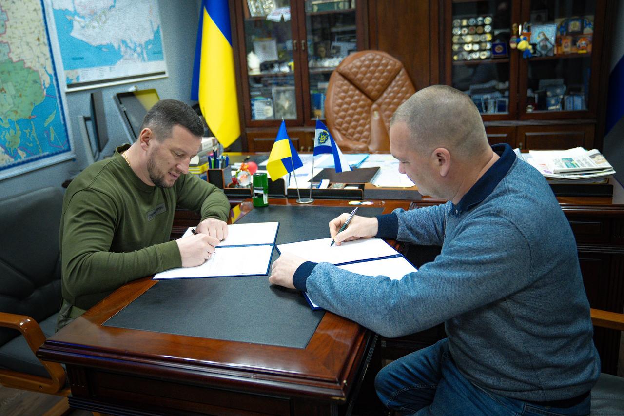 Олександр Прокудін та Дмитро Зорик сидять за робочим столом у кабінеті й одночасно підписують меморандум про співпрацю у сфері кібербезпеки між Херсонською ОВА та Держспецзв’язком.
