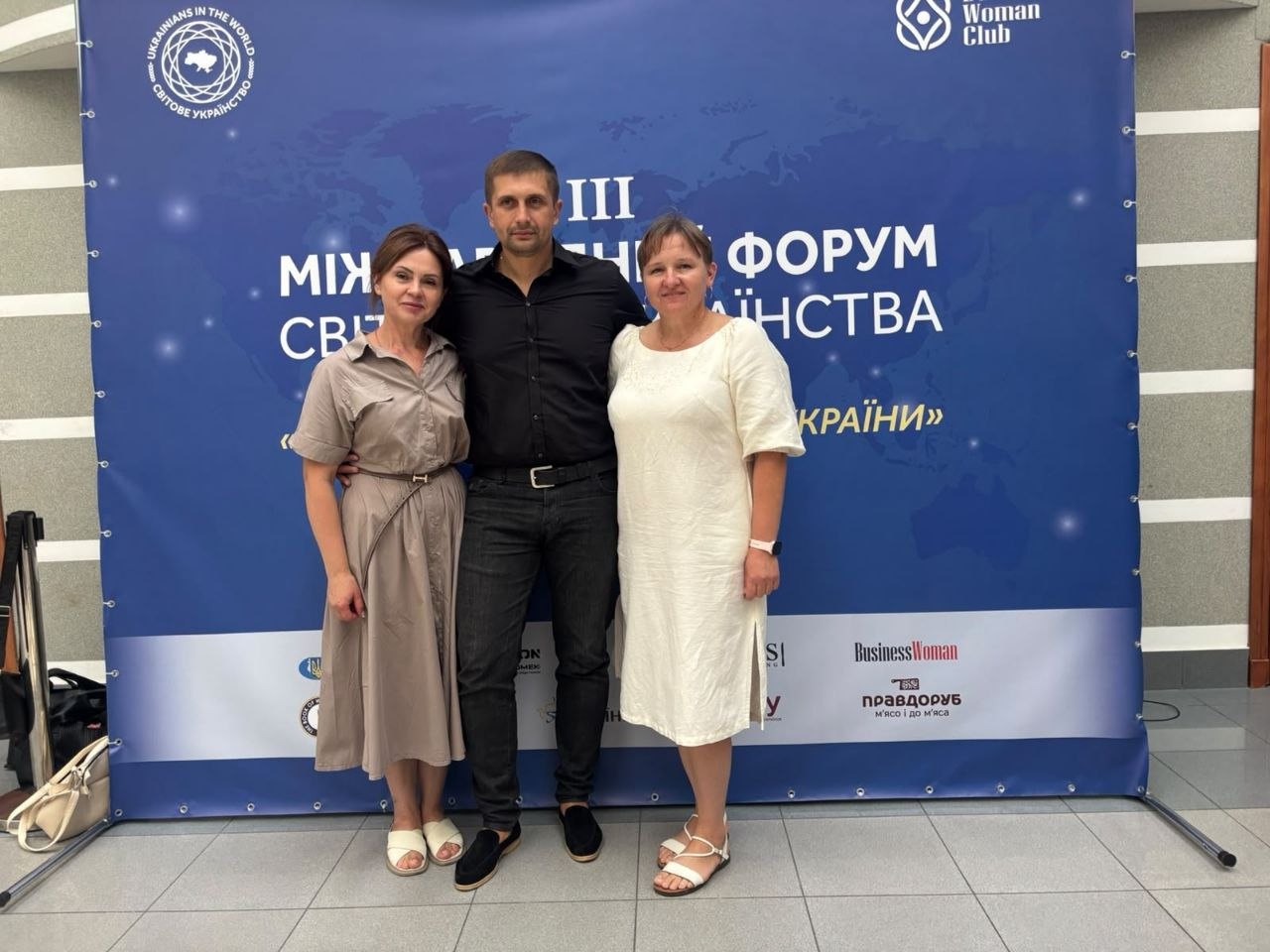 Троє учасників офіційного заходу позують для фотографії перед темно-синім банером-фотозоною III Міжнародного форуму Світового українства.  У центрі стоїть чоловік у чорній сорочці з розстебнутим коміром, темних джинсах та чорному взутті, обіймаючи за плечі жінок з обох боків. Ліворуч стоїть жінка в сіро-бежевій сукні міді з поясом та світлих босоніжках. Праворуч — жінка у білій сукні прямого крою та білих босоніжках.