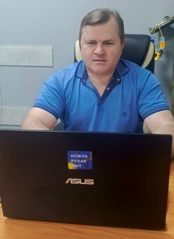 Заступник начальника Херсонської ОВА Володимир Клюцевський у блакитній футболці-поло сидить за робочим столом і зосереджено дивиться в монітор чорного ноутбука Asus. На кришці ноутбука наклеєно синьо-жовту стікер-наліпку з написом «ОСВІТА РУХАЄ СВІТ». На задньому плані видно частину робочого кабінету, крісло та патріотичний сувенір із соняшником і стрічками.