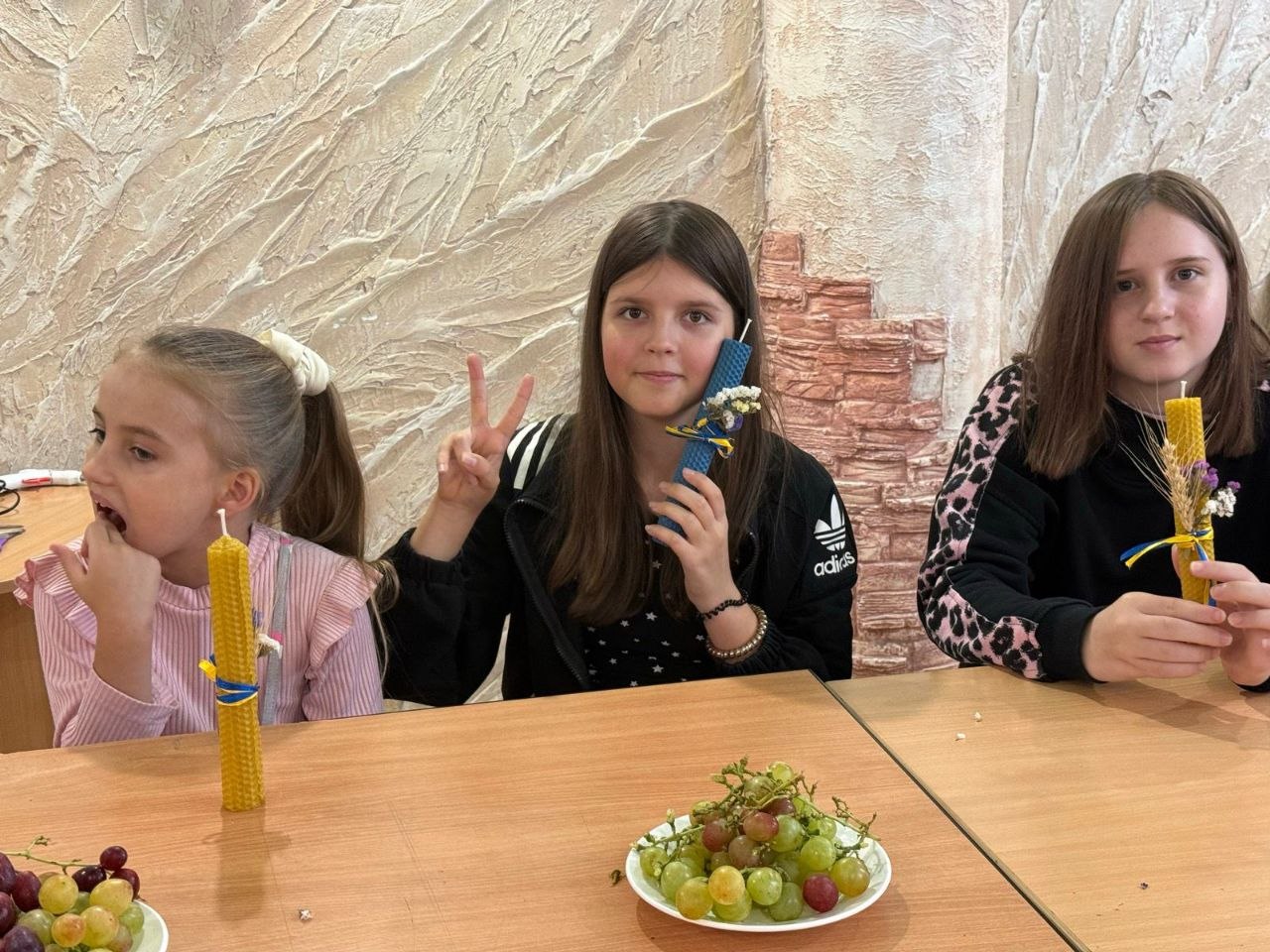 Троє усміхнених дівчат сидять за столом та демонструють зроблені власноруч свічки з натуральної вощини синього й жовтого кольорів, прикрашені колосками та синьо-жовтими стрічками. Дівчинка по центру показує рукою знак «мир» (V). На столі перед ними стоїть тарілка з гронами свіжого винограду.