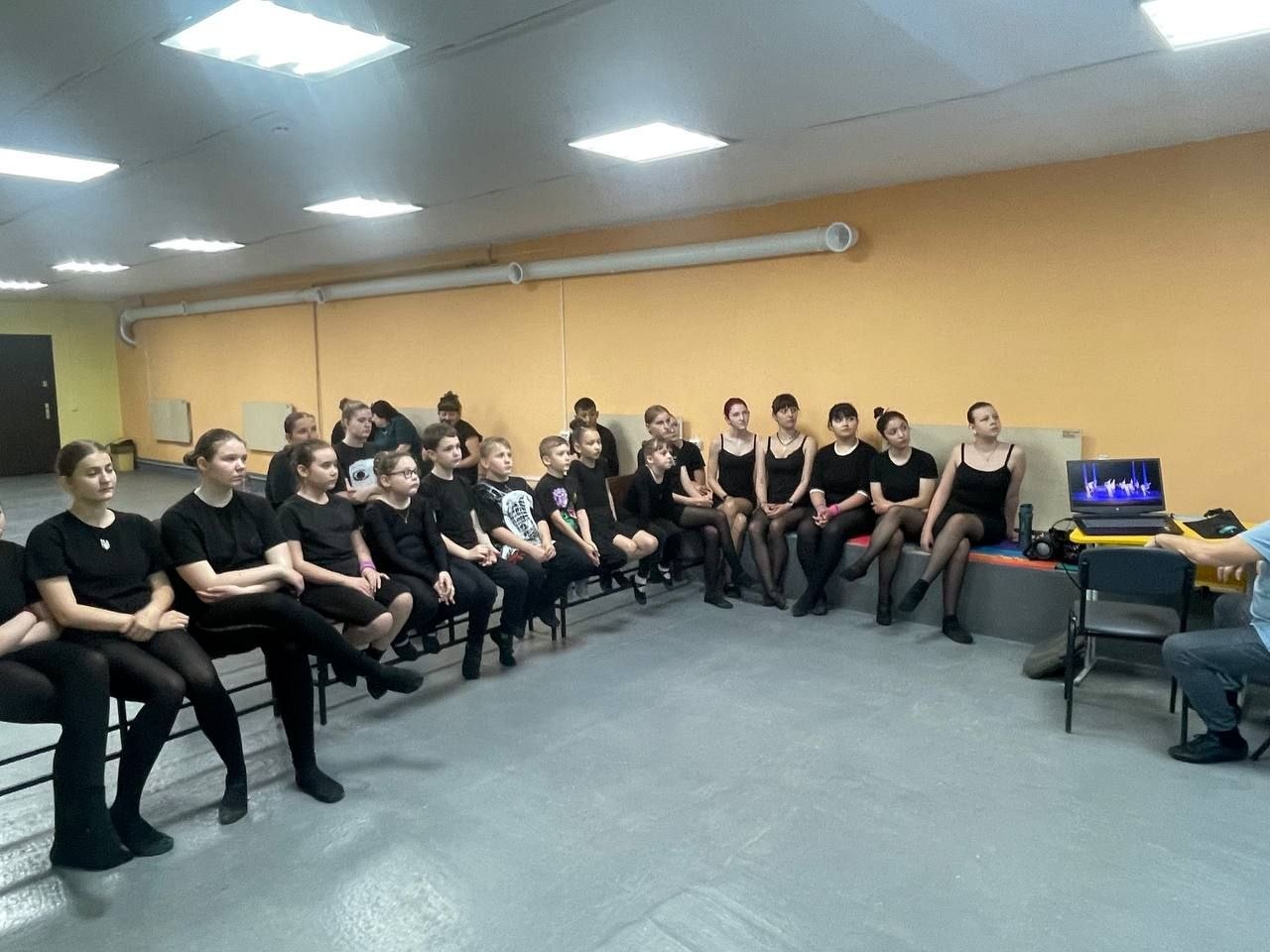 Танцювальний клас із жовтими стінами. Велика група дітей та підлітків (близько 20 осіб) у чорному тренувальному одязі сидить на лавах та матах уздовж стіни. Усі вони дивляться в один бік — на екран ноутбука, що стоїть на столі праворуч. Перед ноутбуком спиною до камери сидить чоловік, який демонструє відеозапис хореографічної постановки на синьому тлі.