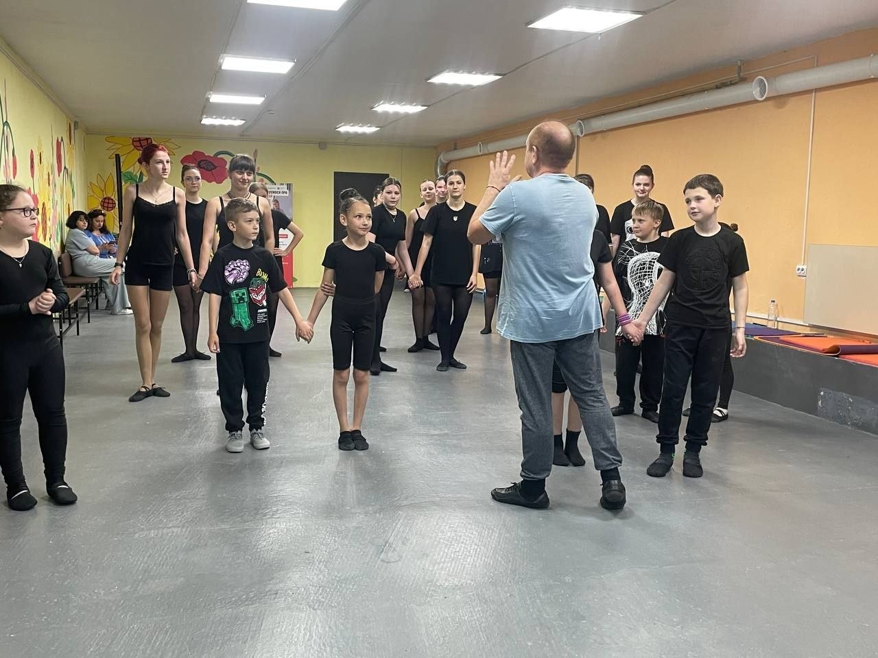 Процес репетиції у танцювальному класі з сірою підлогою та світлими стінами. По центру спиною до камери стоїть чоловік-викладач у блакитній футболці та сірих спортивних штанях, який жестикулює руками, пояснюючи рухи. Перед ним півколом стоять вихованці різного віку (діти та підлітки) у чорній тренувальній формі. На передньому плані хлопчик і дівчинка тримаються за руки, уважно слухаючи вчителя. На задньому плані видно малюнки на стіні.