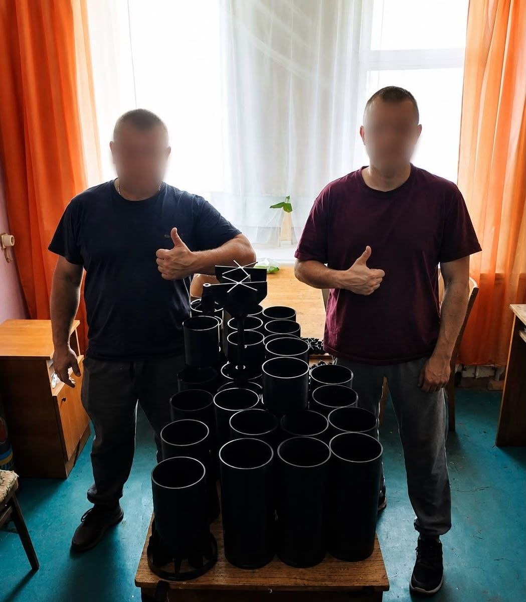 Двоє чоловіків із розмитими обличчями стоять по обидва боки від невисокого столу на тлі вікна з помаранчевими шторами, демонструючи жест схвалення. На столі пірамідою вишикувана велика партія надрукованих на 3D-принтері чорних пластикових корпусів і стабілізаторів для боєприпасів до БпЛА.