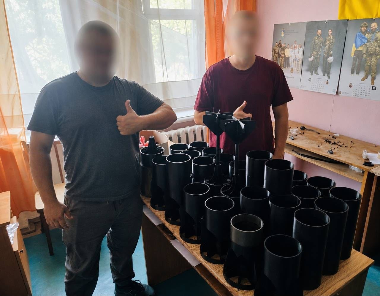 Двоє волонтерів у цивільному одязі з розмитими для безпеки обличчями стоять біля дерев'яного столу в майстерні та показують жест «великий палець угору». На столі щільними рядами розставлені готові пластикові вироби чорного кольору для безпілотників — циліндричні корпуси та хвостові стабілізатори скидів, виготовлені методом 3D-друку. На стіні позаду висять патріотичні календарі із зображеннями українських військових.