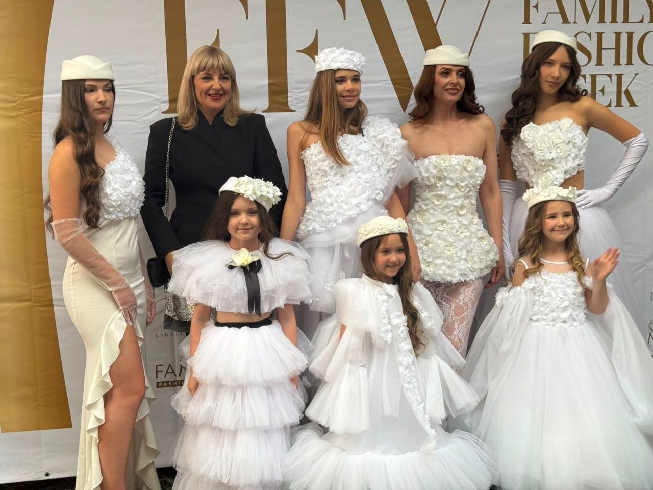 Групове фото з модного показу на тлі світлого банера з написом «FAMILY FASHION WEEK». У центрі та на передньому плані — три маленькі дівчинки у пишних білих фатинових сукнях та капелюшках. Позаду них стоять чотири дорослі моделі в унікальних білих дизайнерських сукнях, прикрашених об'ємними текстильними трояндами, та в білих головних уборах (пілотках і пов'язках). Серед них, у лівій частині кадру, стоїть усміхнена жінка (ймовірно, дизайнерка) у строгому чорному піджаку, що контрастує з рештою образів.