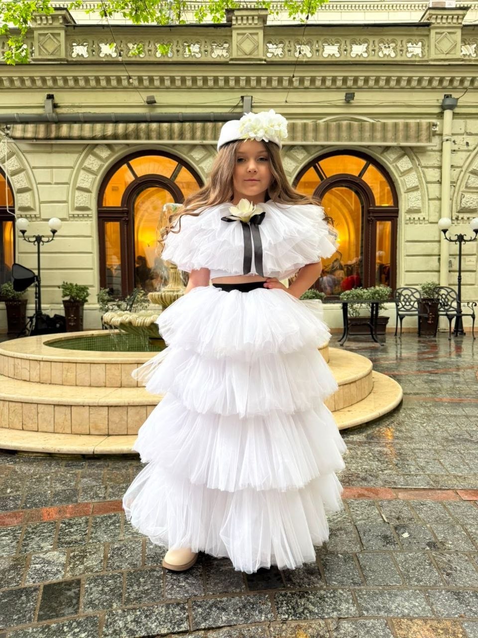 Маленька дівчинка з довгим русявим волоссям позує перед камерою, тримаючи руку на талії. На ній пишний білий костюм: топ із багатошаровими рюшами та великою квіткою з чорною стрічкою по центру, а також довга багатоярусна спідниця з фатину. Образ доповнює біла пілотка, прикрашена дрібними білими квітами. Дівчинка стоїть на мокрій бруківці біля круглого фонтана на тлі архітектурної будівлі з арковими вікнами, що світяться теплим світлом.