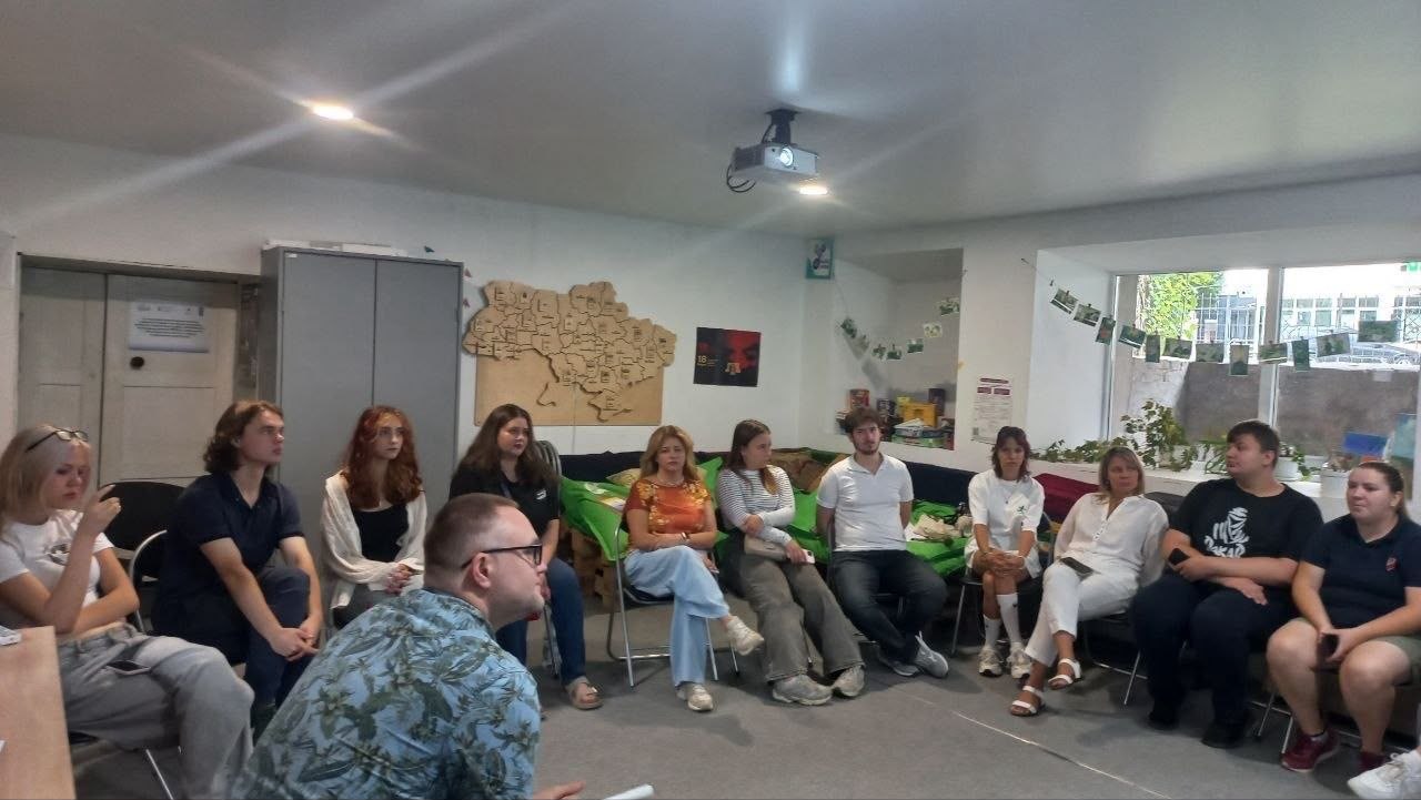 Групова дискусія в просторі молодіжного центру: понад десять учасників різного віку сидять колом уздовж стін кімнати на стільцях та диванах із піддонів. На першому плані спікер веде бесіду з аудиторією. На задній стіні розміщено дерев'яну мапу України з адміністративним поділом областей.