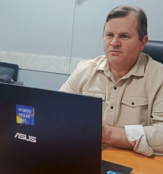 Середнього віку чоловік із темним волоссям, одягнений у бежеву сорочку з накладними кишенями, сидить за дерев'яним столом у кабінеті й зосереджено дивиться на екран ноутбука. На передньому плані видно задню панель чорного ноутбука бренду «ASUS», на яку наклеєно невеликий патріотичний стікер у синьо-жовтих кольорах із написом «ОСВІТА РУХАЄ СВІТ». Чоловік сидить в офісному кріслі з високою спинкою на тлі світло-сірої стіни.