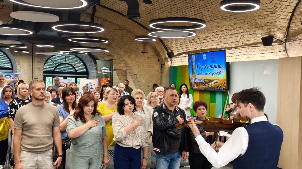 Урочисте відкриття заходу в просторій залі із цегляними склепінчастими арками та кільцевими підвісними світильниками. На першому плані зі спини скрипаль у білій сорочці та синій жилетці грає на скрипці. Перед ним стоїть чисельна аудиторія, люди тримають праву руку на серці під час звучання Державного Гімну України. Праворуч на стіні встановлено телевізійний екран із заставкою «Херсонщина – символ нескореності».