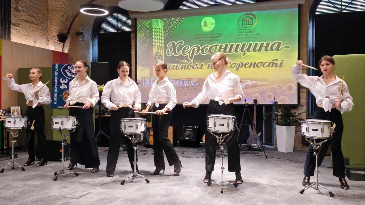 Хореографічно-музичний перформанс ансамблю барабанщиць на сцені. П'ятеро дівчат у білих сорочках, зав'язаних на талії, та широких чорних штанах синхронно грають паличками на маршових барабанах, встановлених на стійках перед екраном із логотипами Департаменту реалізації гуманітарної політики Херсонської ОДА та Херсонського обласного палацу культури.