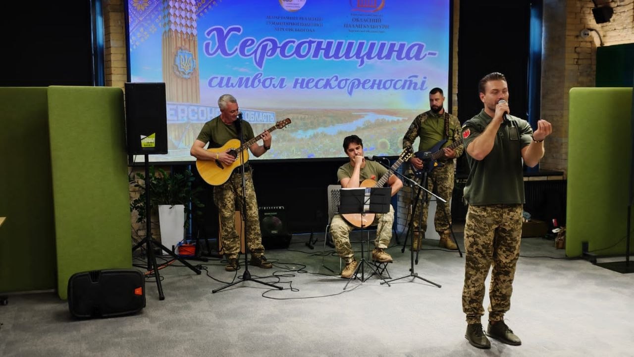 Музичний виступ військового ансамблю на сцені перед екраном «Херсонщина – символ нескореності». На передньому плані соліст у камуфляжних штанах та футболці кольору хакі співає в мікрофон. Позаду нього троє військовослужбовців акомпанують на акустичній гітарі, електрогітарі та бас-гітарі.