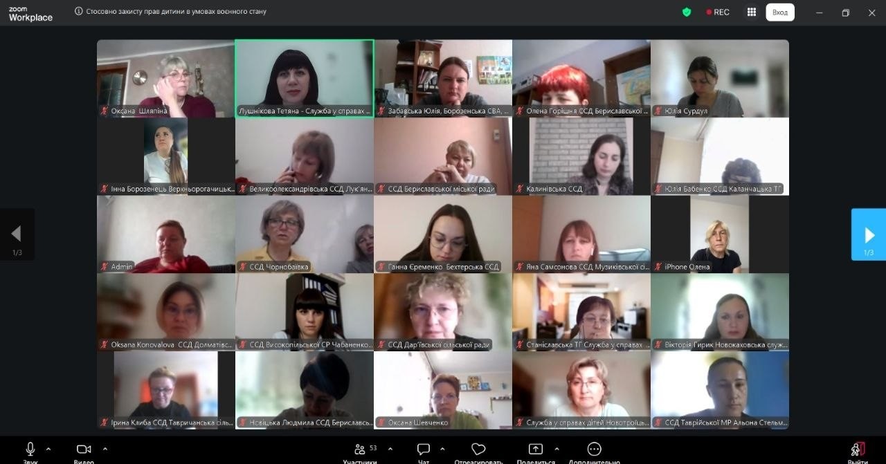 Скриншот онлайн-наради в додатку Zoom Workplace, інтерфейс якого відображає сітку з вікон відеозв'язку учасників (п'ять рядів по п'ять вікон у кожному). У засіданні беруть участь представники служб у справах дітей (ССД) та територіальних громад Херсонщини. У верхній лівій панелі додатку вказано тему зустрічі: «Стосовно захисту прав дитини в умовах воєнного стану». У верхньому правому кутку горить червоний індикатор запису «REC». Вікно однієї з учасниць у першому ряду підписане як «Лушнікова Тетяна - Служба у справах...» та виділене зеленою рамкою активного спікера.