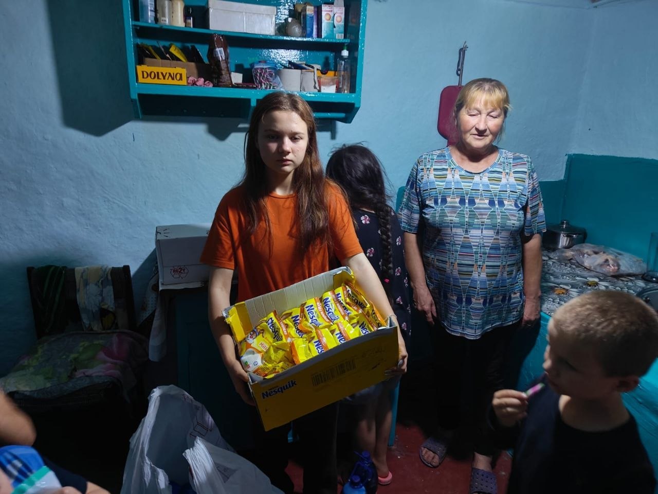 Дівчина в помаранчевій футболці тримає в руках картонну коробку, заповнену пакунками шоколадного напою Nesquik, під час видачі гуманітарної допомоги. Поруч стоять літня жінка у строкатій футболці та діти. На задньому плані видно блакитні стіни кімнати та настінну полицю з побутовими речами й коробками.