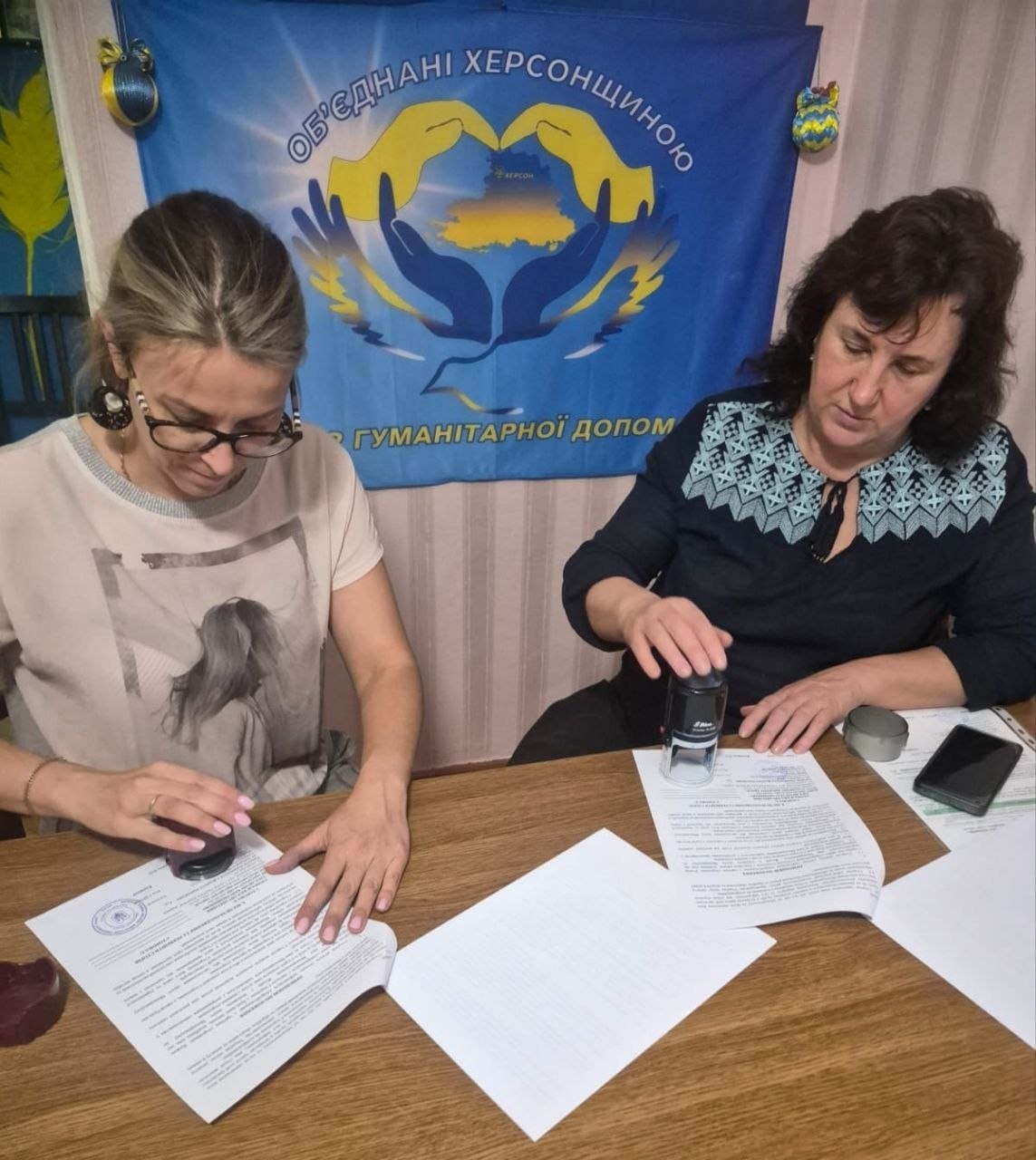 Робочий момент підписання та завірення офіційних документів у хабі допомоги. Дві жінки сидять за дерев'яним столом і ставлять печатки на аркуші угод. Позаду них на стіні закріплено синій прапор із написом «ОБ'ЄДНАНІ ХЕРСОНЩИНОЮ», емблемою долонь, які тримають карту області, та написом «ГУМАНІТАРНОЇ ДОПОМОГИ».
