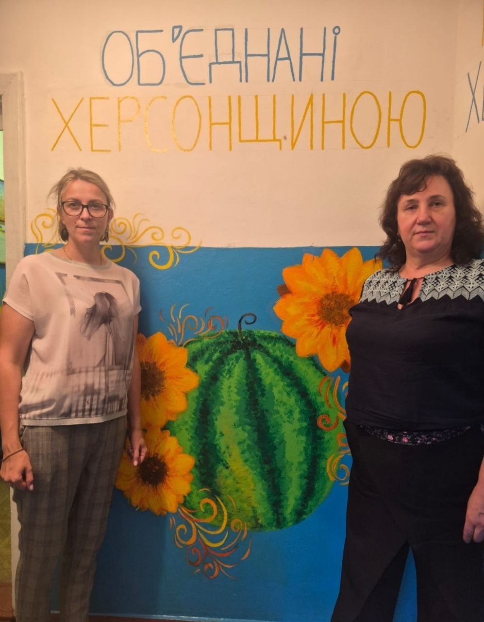 Дві жінки стоять біля розписаної стіни гуманітарного простору. На стіні зверху нанесено напис великими літерами «ОБ'ЄДНАНІ ХЕРСОНЩИНОЮ», а нижче зображено яскравий стінопис із великим смугастим кавуном в оточенні соняшників та декоративних візерунків на синьому тлі.