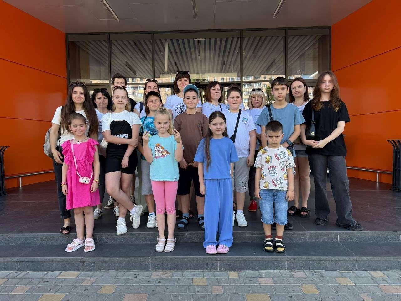 Спільне групове фото дітей та дорослих (близько двадцяти учасників) на подвір'ї перед входом до сучасної будівлі. Група вишикувалася у кілька рядів на гранітних сходах.  На передньому плані стоять четверо дітей:  крайня ліворуч — дівчинка в яскраво-рожевій літній сукні з невеликою білою сумочкою через плече;  поруч із нею — дівчинка у світло-блакитній футболці та рожевих лосинах;  далі стоїть дівчинка у довгому вільному блакитному костюмі;  крайній праворуч — маленький хлопчик у білій футболці з яскравими принтами та джинсових шортах.  Позаду них стоять інші діти, підлітки та дорослі жінки. Усі присутні дивляться в об'єктив камери. Будівля на задньому плані має масивні скляні двері, вітринні вікна та яскраво-помаранчеве оздоблення стін. Перед сходами укладено сіро-жовту тротуарну бруківку.