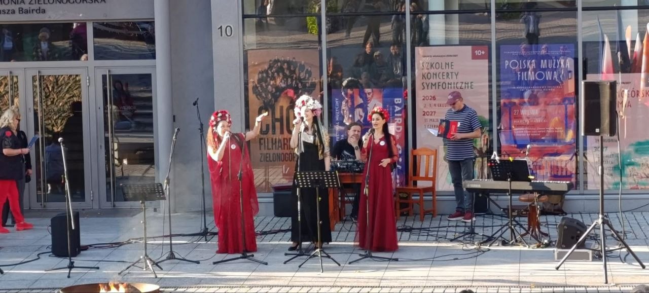 Вуличний виступ вокального колективу біля будівлі Зеленогурської філармонії (Польща). Троє солісток у традиційних українських вінках — двоє в довгих червоних сукнях та одна в чорному вбранні з білою блузою — співають у мікрофони перед пюпітрами. Праворуч за синтезатором стоїть музикант, поруч розташований ведучий у кепці з папкою для виступів, а позаду на вітринах філармонії видно афіші подій.