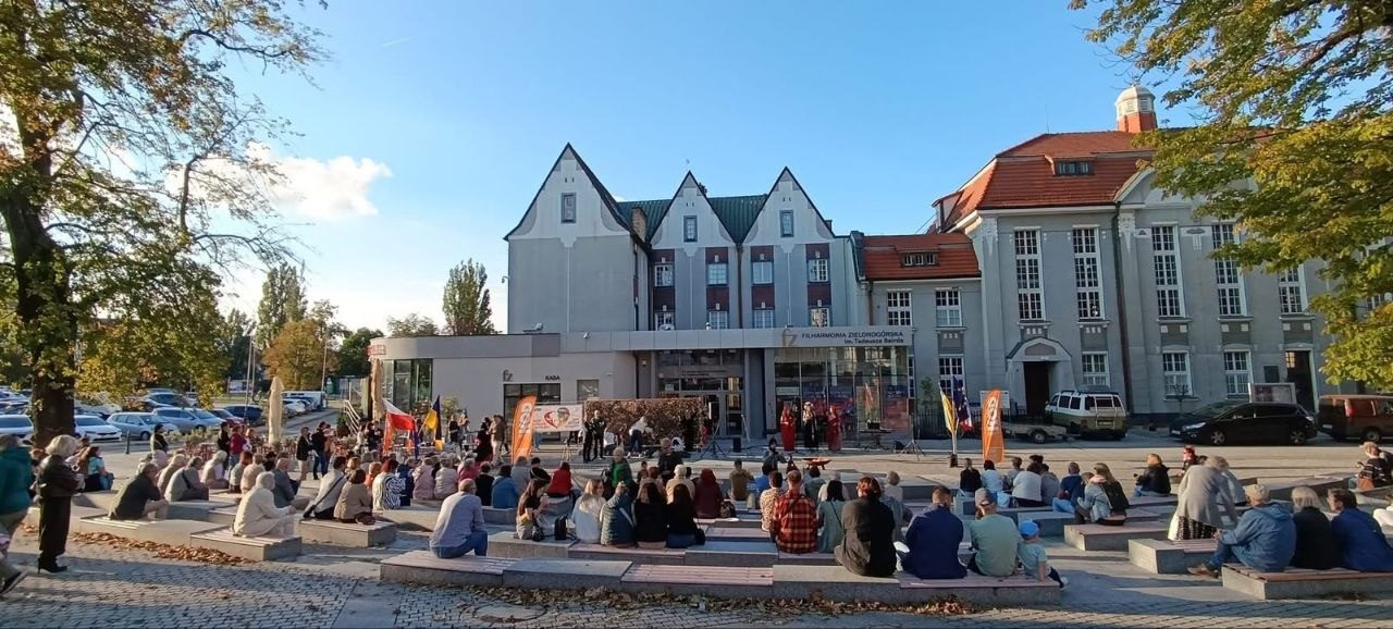 Загальний план площі перед сучасною будівлею філармонії в Зеленій Гурі під час культурно-просвітницької акції. Численні глядачі сидять ярусами на сходах та лавах вуличного амфітеатру, спостерігаючи за виступом артистів на відкритій сцені біля входу. Навколо площі ростуть зелені дерева та припарковані авто