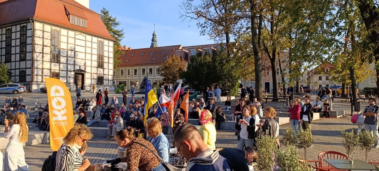 Громадський захід на площі європейського міста в сонячний день. На передньому плані волонтери роздають частування та спілкуються біля жовтого промостенду «KOD». У центрі майдану встановлено прапори України та Польщі, а на кам'яних лавах амфітеатру розмістилися десятки містян. На тлі видно історичну фахверкову будівлю та міську ратушу зі шпилем