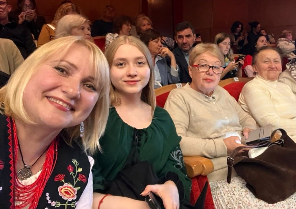 Селфі чотирьох глядачок у глядацькій залі перед виставою. На передньому плані усміхається білява жінка в чорному вишитому кептарі з червоним намистом, поруч сидить юна дівчина в смарагдовій блузі, а далі у ряду — дві літні пані на м'яких червоних кріслах театру. На задньому плані видно інших глядачів.