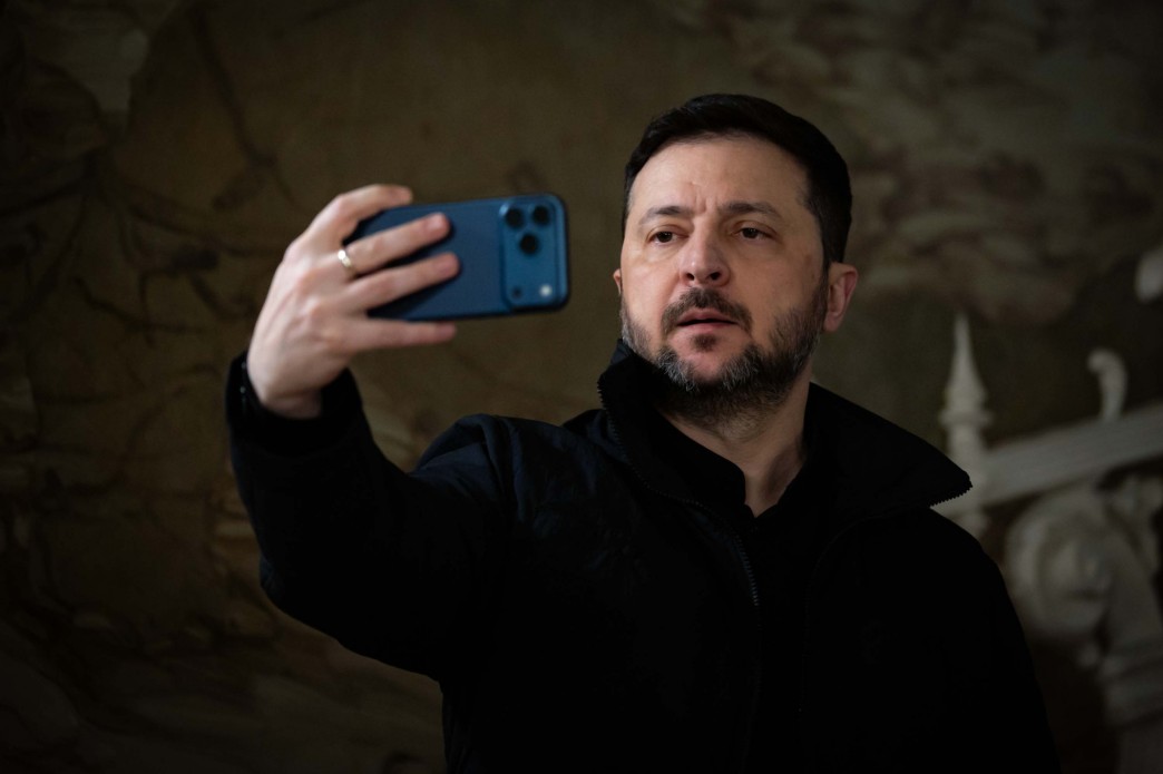 Чоловік у темному одязі робить селфі або веде відеозйомку на смартфон. Він тримає телефон у лівій руці на рівні обличчя, дивлячись в екран. Зйомка відбувається у приміщенні з приглушеним світлом.