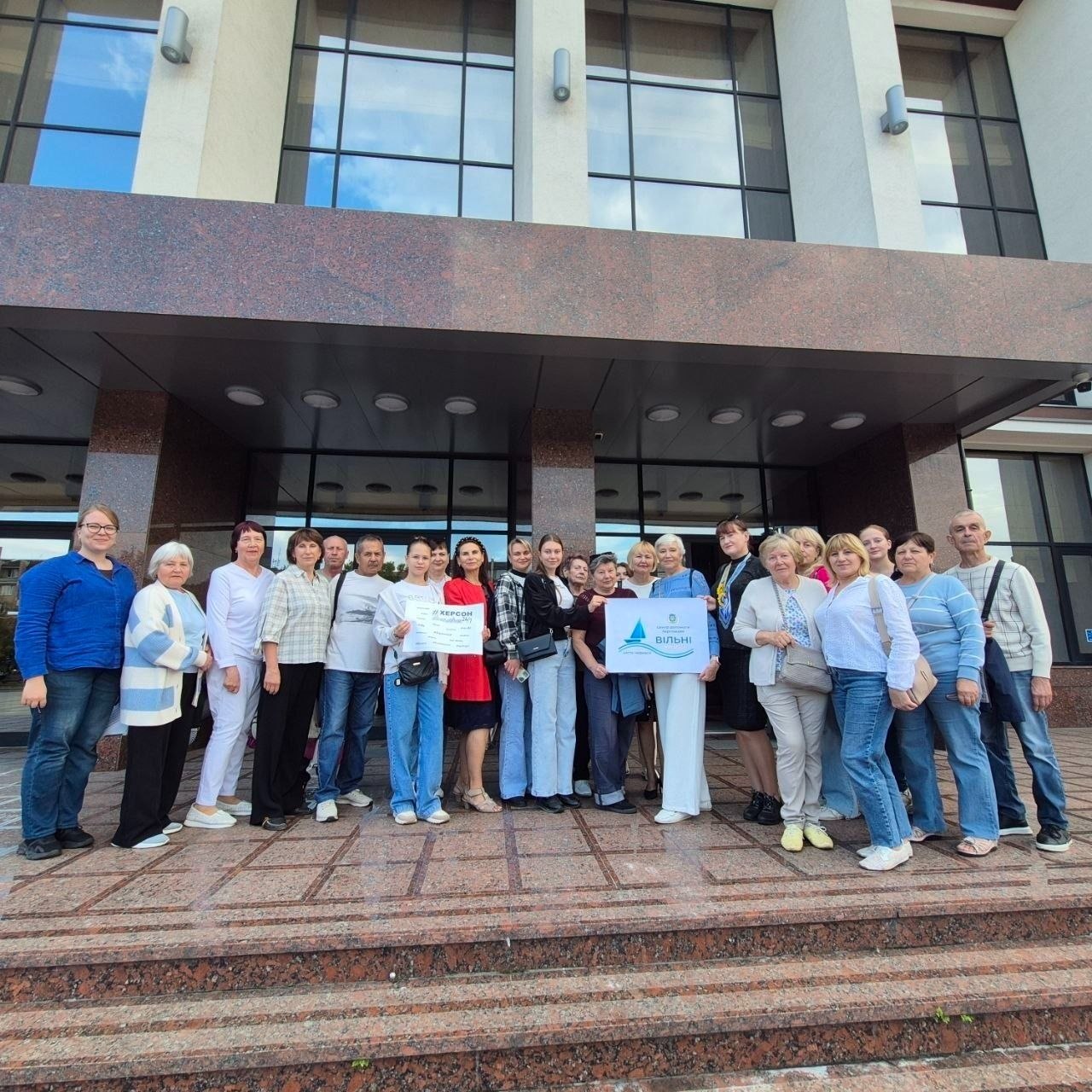 Велика група людей різного віку позує на широких кам'яних сходах перед сучасним входом до театру або палацу культури з високими колонами та темними вітражними вікнами. У центрі учасники тримають у руках таблички: невелику білу з написом «#ХЕРСОН» та більшу горизонтальну з логотипом простору «ВІЛЬНІ».