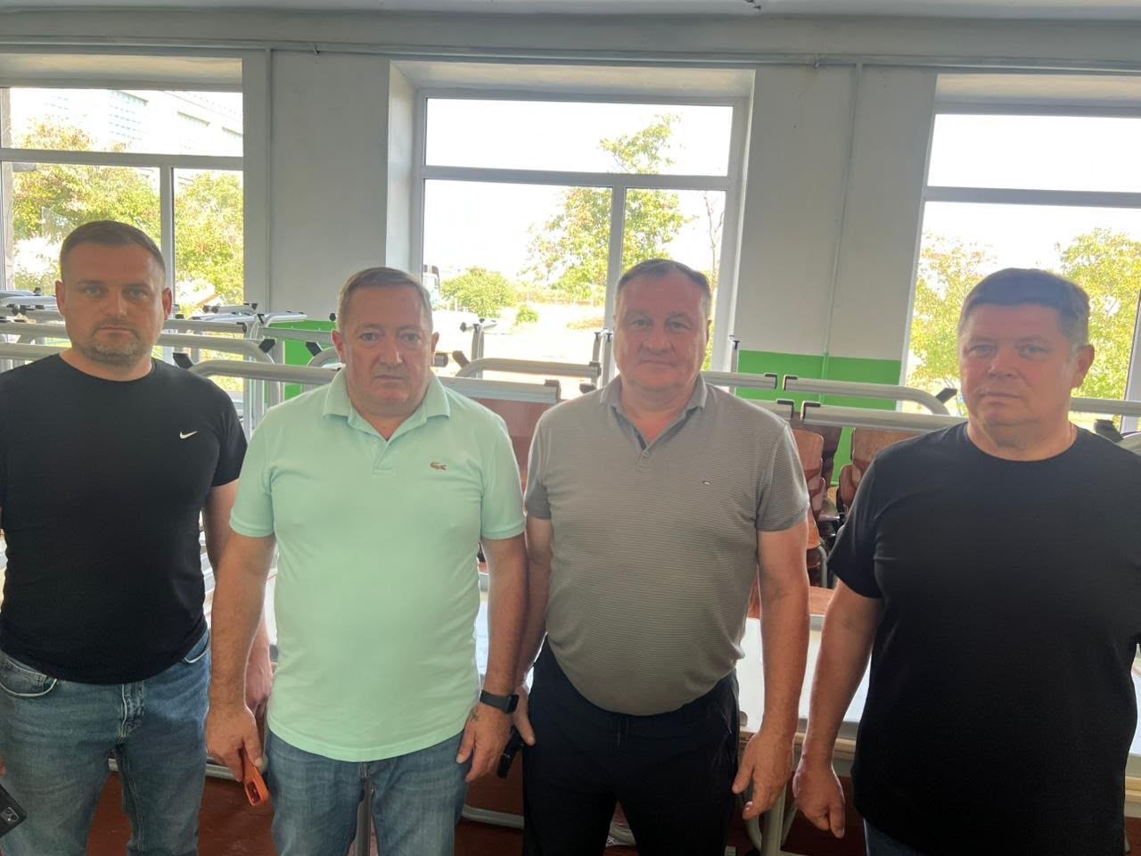 Четверо чоловіків стоять у шкільному приміщенні на тлі великих вікон та нових металевих конструкцій учнівських парт. Вони позують для фотографії, оглядаючи облаштування та оновлення навчального простору