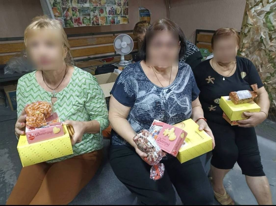 Троє жінок тримають перед собою подарункові кондитерські набори: фірмові коробки з німецьким печивом та пакунки із солодощами, зав'язані стрічками. Вони сидять на лаві у волонтерському центрі. На тлі видно робочий простір із вентилятором, коробками та маскувальною сіткою. Обличчя присутніх розмиті.