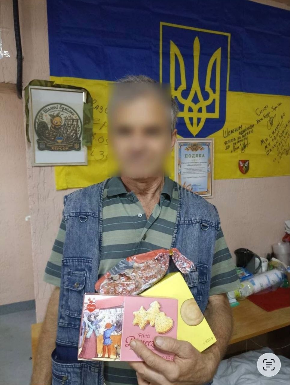 Чоловік у смугастій футболці поло та джинсовому жилеті стоїть у приміщенні та тримає в руках набір кондитерських виробів: рожеву й жовту коробки європейського пісочного печива та прозорий пакунок зі смаколиками. На стіні позаду нього висить синьо-жовтий Державний Прапор України з золотистим Тризубом, підписами військових та подякою, а також рамка з емблемою волонтерської ініціативи «Шалені бджілки. Херсон». Обличчя чоловіка приховане.