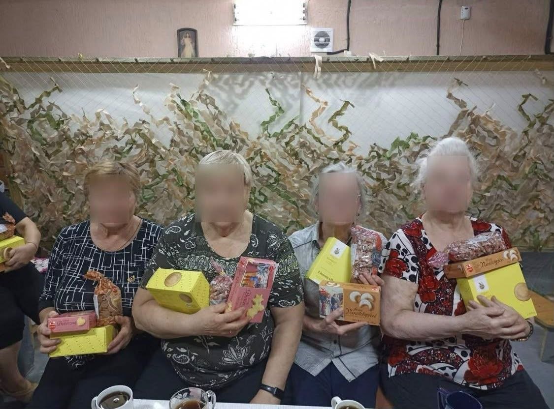 Чотири жінки літнього віку сидять в ряд у приміщенні волонтерського осередку та тримають у руках отримані набори солодких подарунків: жовті та рожеві коробки з печивом, а також прозорі пакунки з солодощами. На передньому плані видно чашки з чаєм або кавою. Позаду на стіні розвішана маскувальна сітка та висить невелика ікона в арковій ніші. Обличчя жінок розмиті для збереження конфіденційності.
