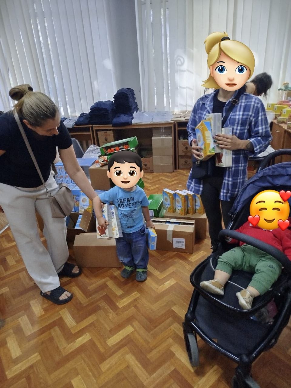 Видача гуманітарної допомоги родинам із дітьми в приміщенні з паркетною підлогою. Маленький хлопчик у блакитній футболці та джинсах тримає пакунки з дитячим харчуванням біля картонних коробок. Поруч стоять жінки, які тримають коробки з допомогою, а праворуч у дитячому візочку сидить немовля. Обличчя дорослих та дітей закриті захисними емодзі.