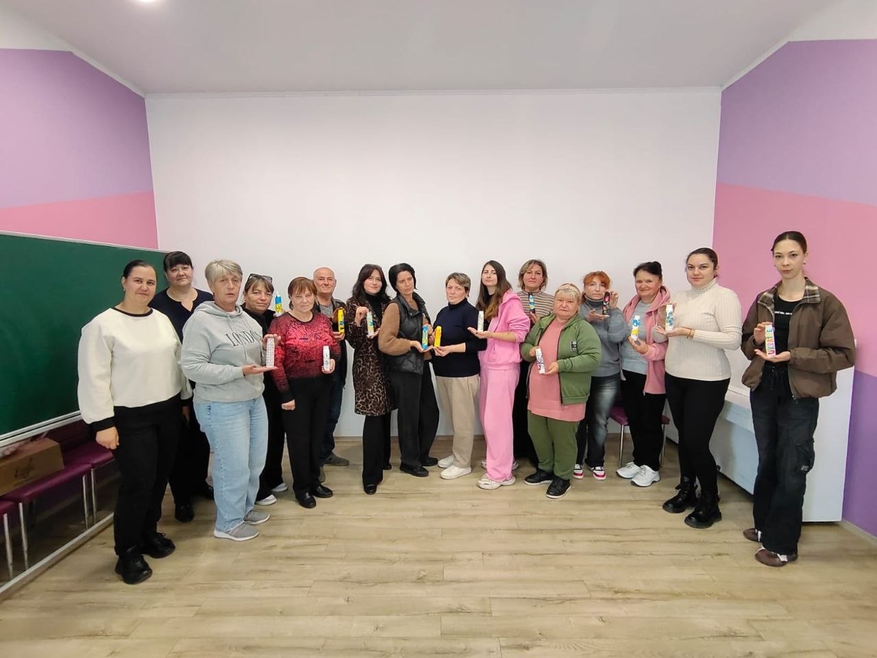 Учасниці та організатори арттерапевтичного майстер-класу вишикувалися для спільної фотографії у просторій світлій кімнаті з бузковими стінами. Жінки та чоловік тримають перед собою власноруч розписані декоративні гільзи й тубуси з квітковими візерунками та українською символікою. Ліворуч біля стіни встановлено шкільну дошку.
