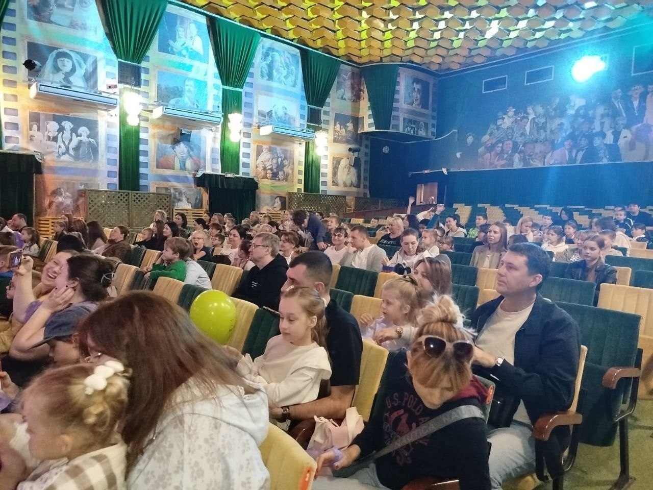 Заповнена глядацька зала театру чи кінотеатру зі стінами, декорованими кінострічками з архівними фотографіями, та зеленою драперією на колонах. Батьки разом із дітьми сидять у рядах крісел, очікуючи на виступ артистів. На передньому плані дівчинка тримає салатову повітряну кульку, глядачі спілкуються та фотографують сцену на смартфони.