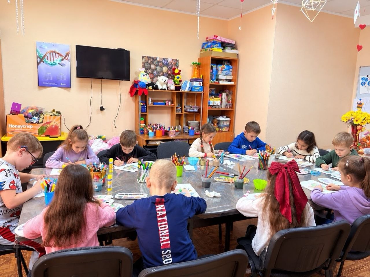 Група дітей молодшого віку сидить за спільним великим столом під час арттерапевтичного заняття у світлій ігровій кімнаті. Діти розфарбовують малюнки олівцями та фломастерами. На задньому плані видно полиці з м'якими іграшками, розвивальними настільними іграми, телевізор на стіні та підвісні декоративні сердечка під стелею.