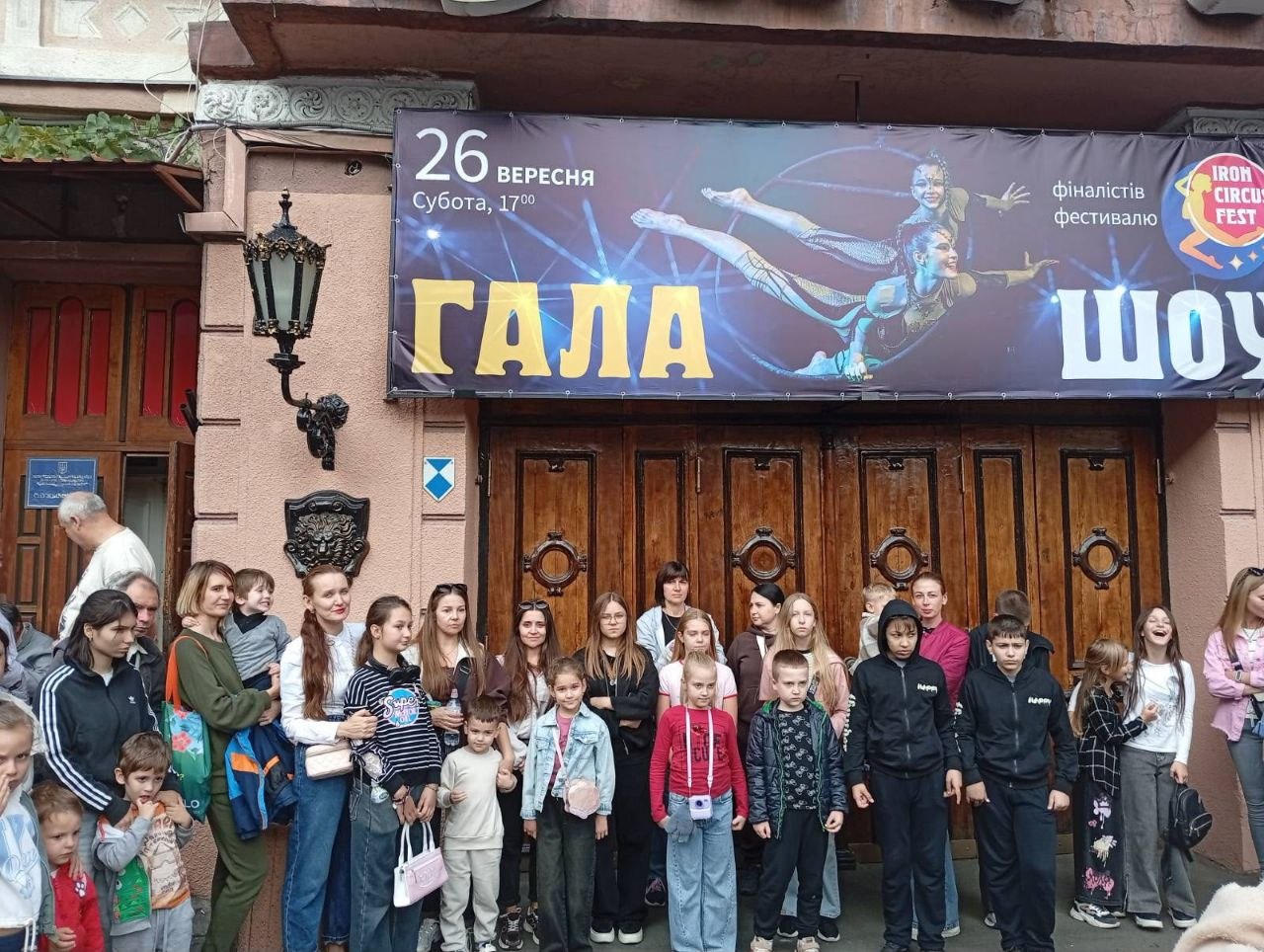 Велика група дітей, підлітків та їхніх батьків стоїть біля масивних дерев'яних різьблених дверей цирку перед початком вистави. Над входом висить великий темний рекламний банер заходу «ГАЛА ШОУ» з фотографією гімнастки в польоті, логотипом «Iron Circus Fest» і датою: «26 вересня, субота, 17:00». Зліва на фасаді розташований кований ліхтар та барельєф із головою лева.