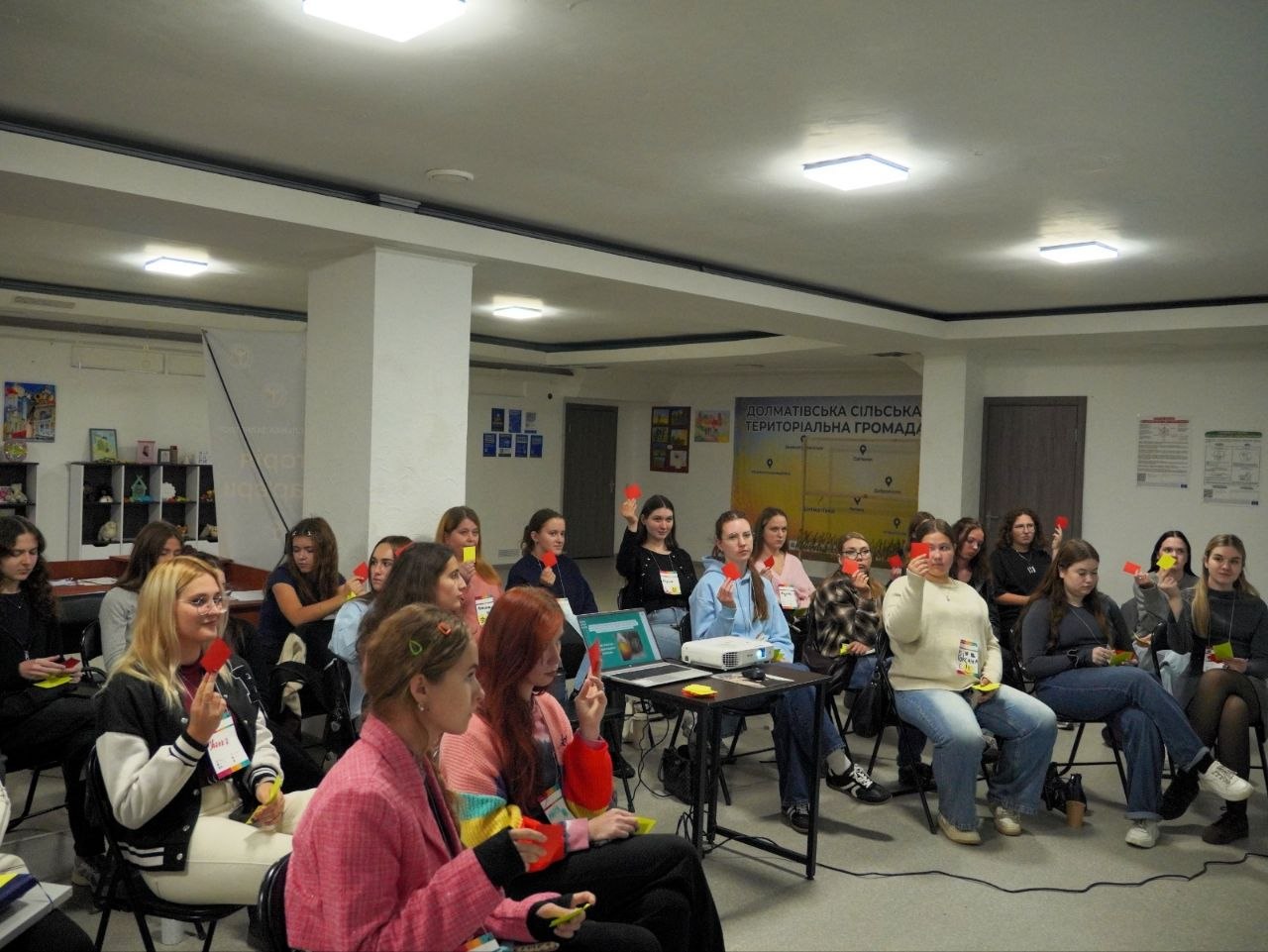 Практичне інтерактивне опитування в аудиторії навчального центру. Дівчата та молоді жінки сидять на стільцях перед проєктором і ноутбуком, тримаючи в руках та піднімаючи вгору кольорові сигнальні картки червоного й жовтого кольорів для голосування. На задньому плані розташовано інформаційний стенд Долматівської сільської територіальної громади