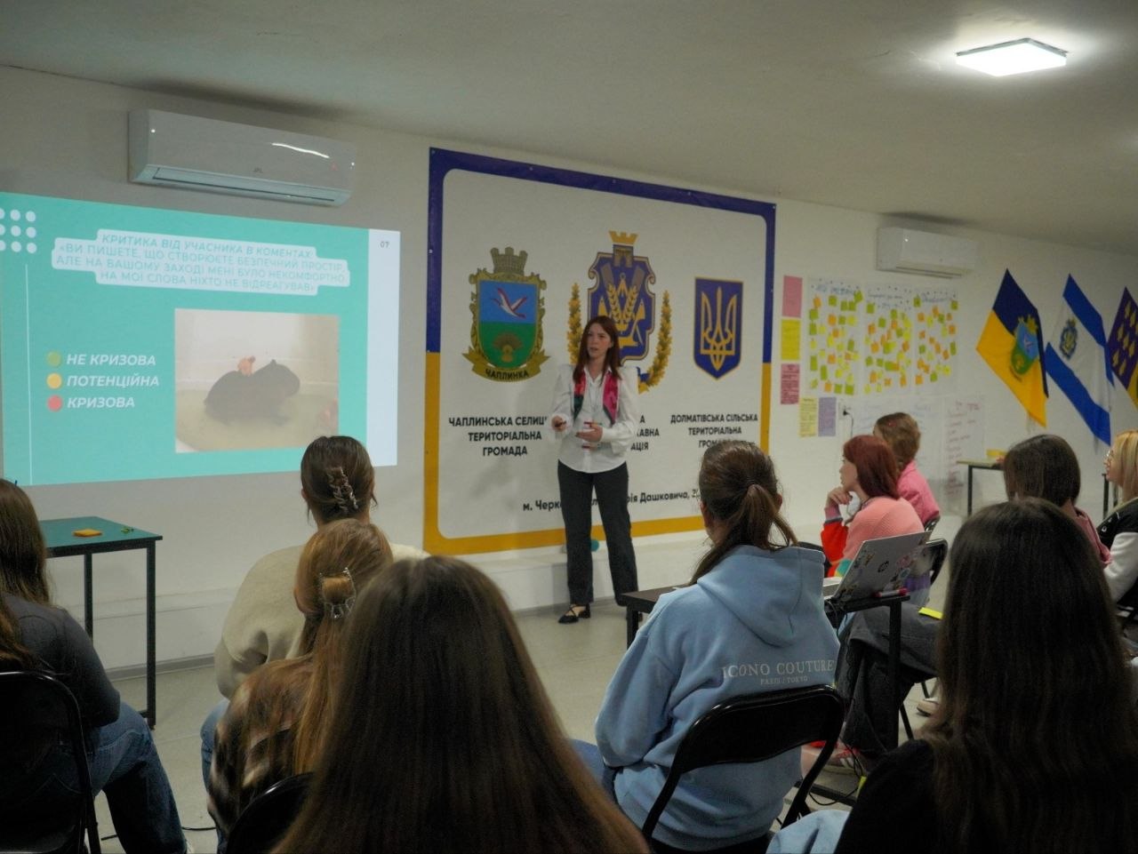 Лекторка стоїть перед аудиторією біля демонстраційного екрана та брендволу з державною й регіональною символікою Херсонщини. На слайді презентації розглядається тема «Критика від учасника в коментарях» із класифікацією: не кризова, потенційна та кризова ситуація. Учасниці сидять спиною до камери та слухають лекцію, одна з них працює за ноутбуком