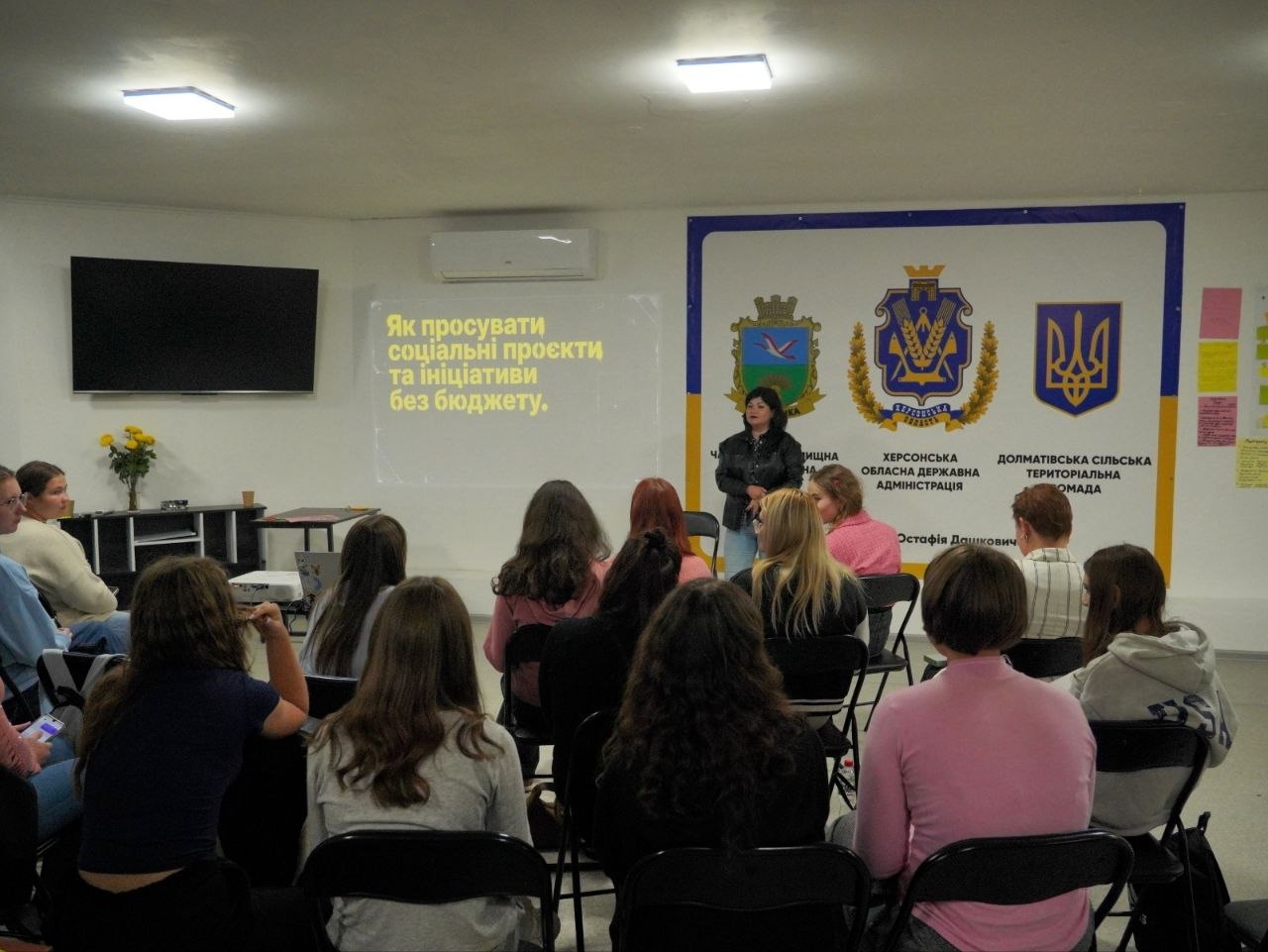 Вид із задніх рядів навчальної аудиторії на виступ тренерки. На проєкційному екрані ліворуч відображено назву модуля: «Як просувати соціальні проєкти та ініціативи без бюджету». Праворуч за лекторкою розміщено банер із гербами України, Херсонської ОДА, Чаплинської та Долматівської громад. Слухачки зосереджено дивляться на спікерку