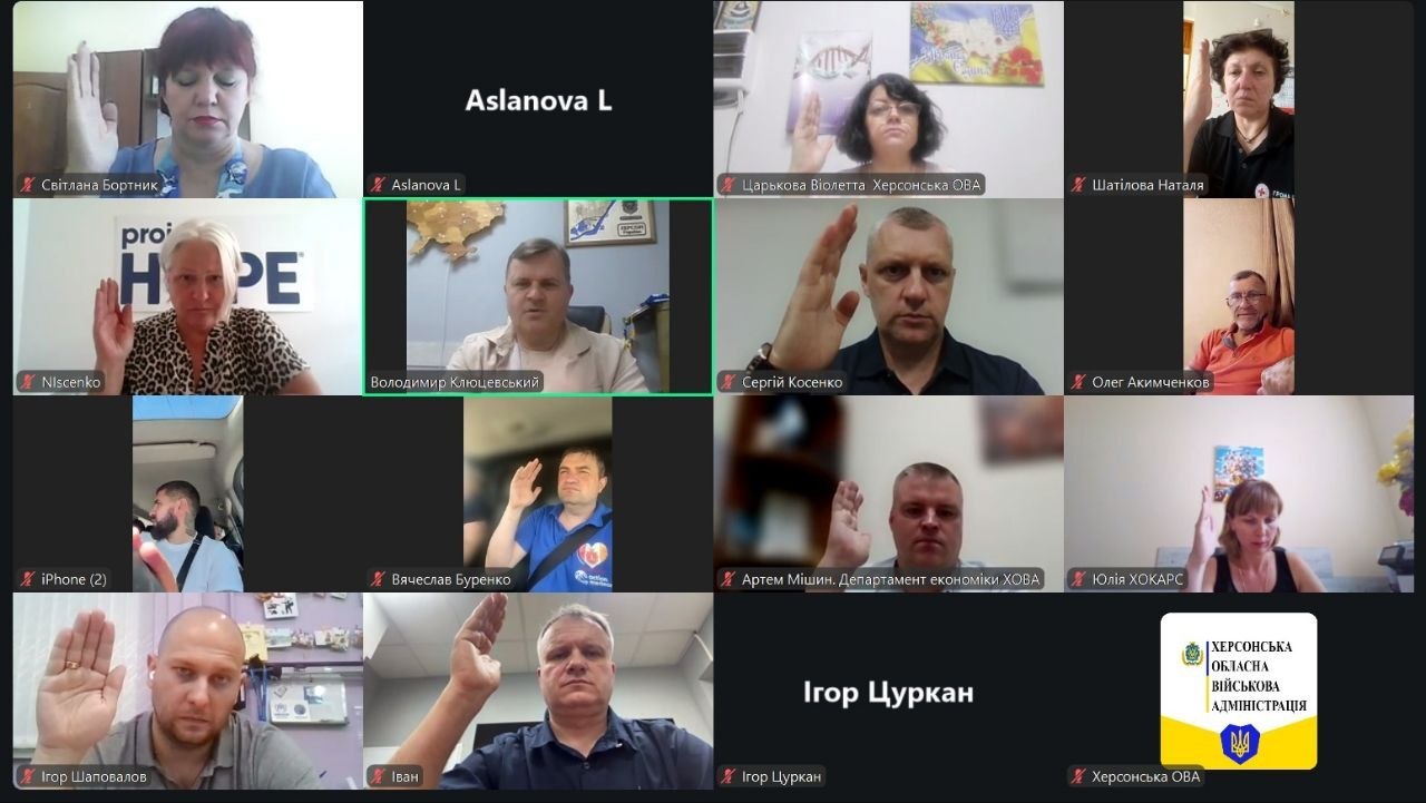 Скриншот екрана під час проведення робочої наради чи засідання у форматі онлайн-відеоконференції на платформі Zoom. Учасники заходу — представники Херсонської обласної військової адміністрації (ХОВА) та партнерських організацій.  Відеочат розділений на сітку з 14 вікон, де більшість присутніх одночасно підняли праву руку вгору, беручи участь у процедурі голосування.  Серед вікон конференції зафіксовані:  По центру у другому ряду (виділений зеленою рамкою активного промовця) — заступник начальника Херсонської ОВА Володимир Клюцевський, який сидіти у робочому кабінеті на тлі мапи України.  Також увімкнені відеотрансляції інших посадовців та учасників із підписами: Світлана Бортник, Віолетта Царькова (Херсонська ОВА), Наталя Шатілова, Сергій Косенко, Олег Акимченков, Вячеслав Буренко, Артем Мішин (Департамент економіки ХОВА), Юлія ХОКАРС, Ігор Шаповалов та Іван.  Одне з вікон ліворуч (підпис «NIsченко») належить представниці благодійної організації: жінка сидить на тлі банера з логотипом «Project HOPE».  Три вікна не мають відеозв'язку і відображають лише текстові імена на чорному тлі: «Aslanova L» та «Ігор Цуркан».  У правому нижньому кутку екрана замість відеоприйому встановлено офіційну заставку-логотип установи: біло-жовтий прямокутник із гербом області, тризубом та написом «ХЕРСОНСЬКА ОБЛАСНА ВІЙСЬКОВА АДМІНІСТРАЦІЯ».
